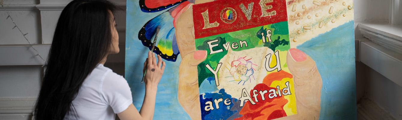 Person sitting and finishing a painting with a blue background, a rainbow coloured butterfly, and a big hand in the middle holding a poster that reads "Love even if you are afraid".