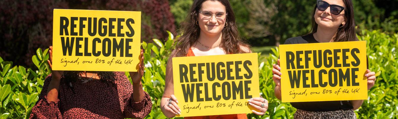 Three women holding a 'Refugees welcome' placard smiling at camera