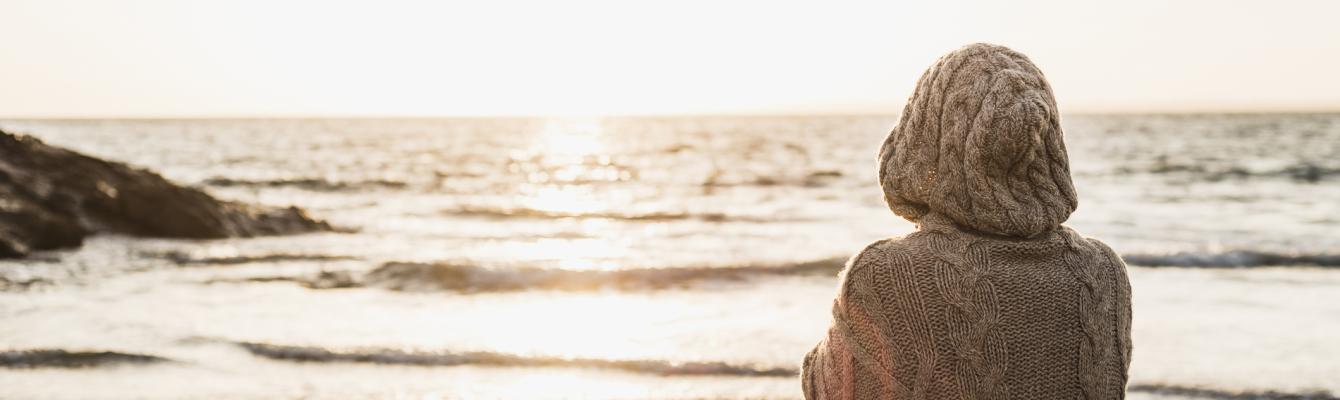 Woman with a hood and back to camera looks onto a beach