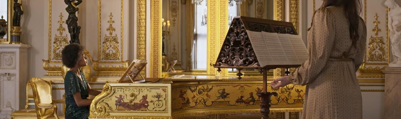 Image of a lady sat playing the piano and a woman opposite singing from a music stand, both in an ornate room.