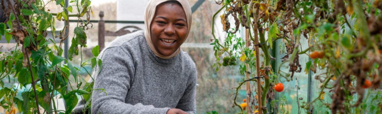 An image of a woman crouching in a greenhouse among vegetable plants, smiling directly at the camera.