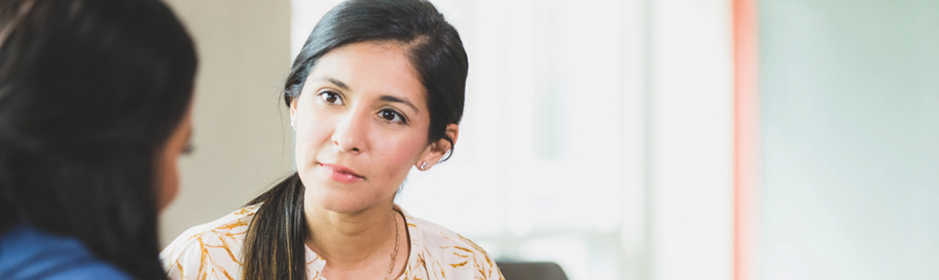 A woman with dark hair in a ponytail listens attentively to another person, sitting indoors in a well-lit space.