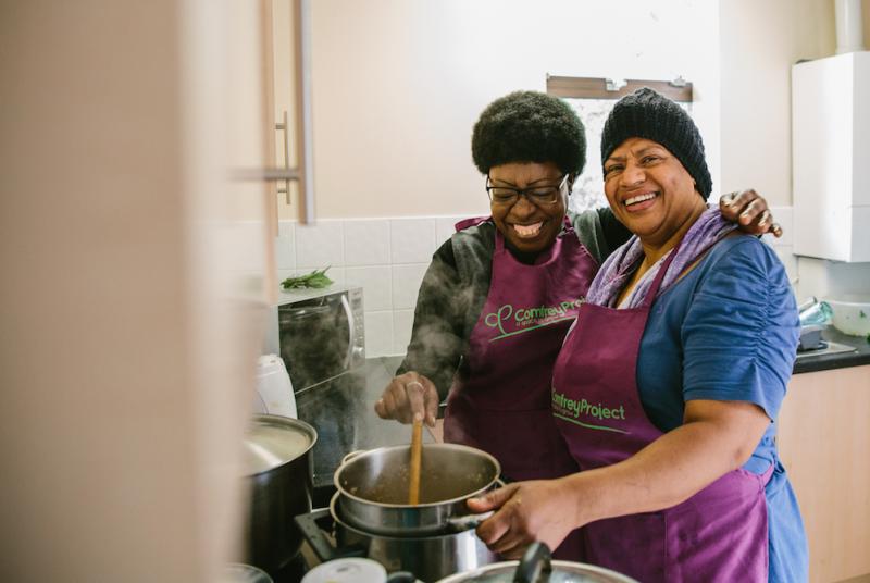 Two smiling women wearing purple Comfrey Project aprons are cooking together 