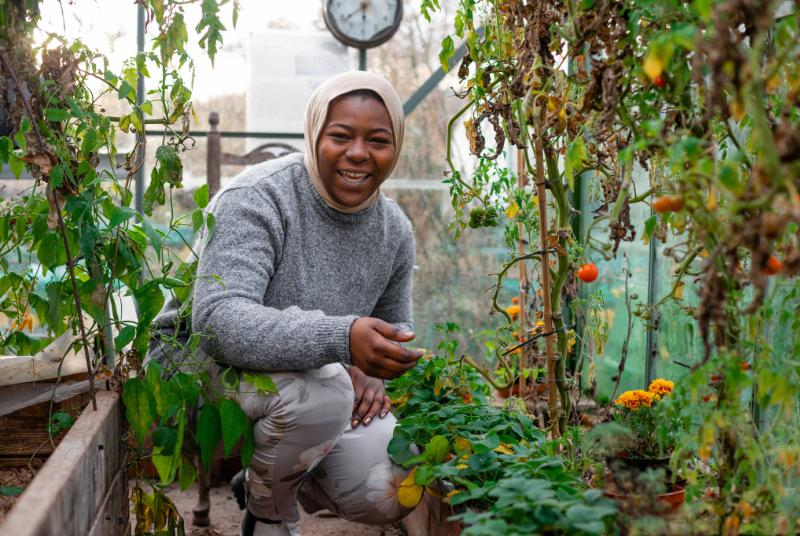 An image of a woman crouching in a greenhouse among vegetable plants, smiling directly at the camera.