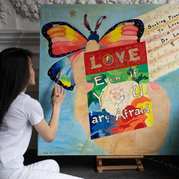 Person sitting and finishing a painting with a blue background, a rainbow coloured butterfly, and a big hand in the middle holding a poster that reads "Love even if you are afraid".