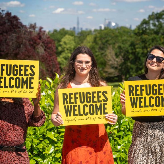 Three women holding a 'Refugees welcome' placard smiling at camera