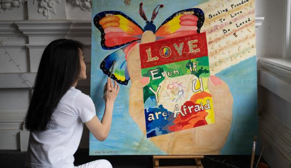 Person sitting and finishing a painting with a blue background, a rainbow coloured butterfly, and a big hand in the middle holding a poster that reads "Love even if you are afraid".
